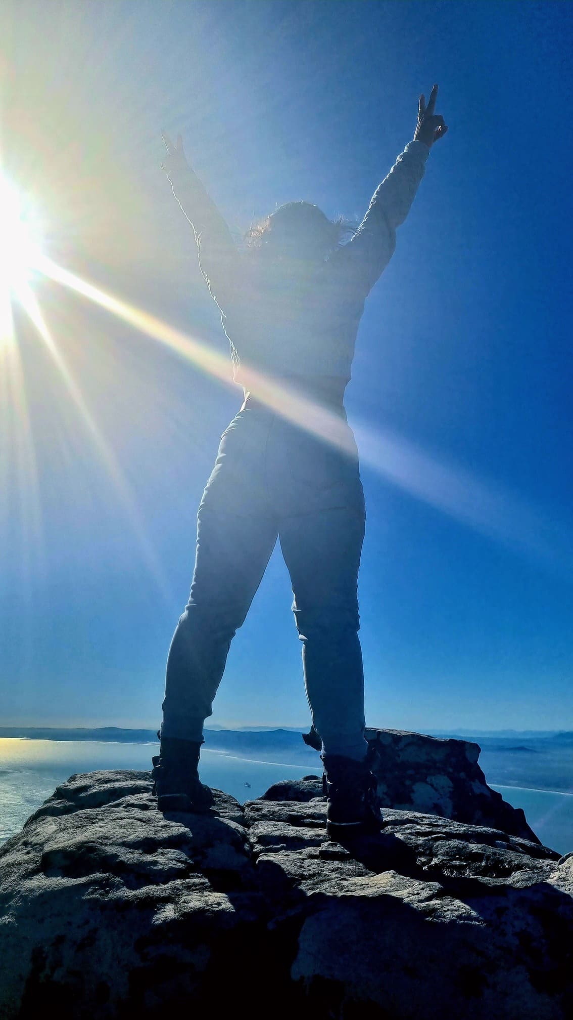 a woman at the top of a mountain celebrating with arms up and sunrays illuminating her victory.